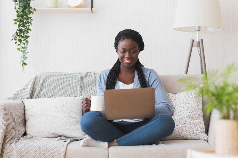 woman blogging on a laptop while sitting cross-legged on couch and holding a cup of coffee